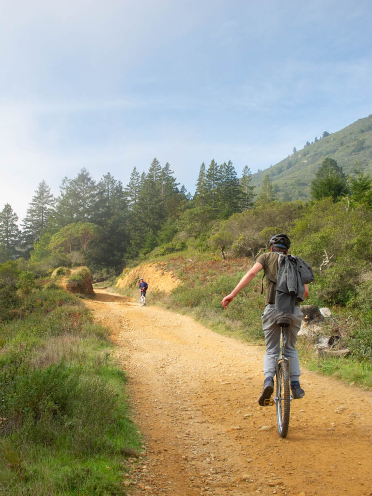 A mountain unicyclist rides away from the camera, climbing a dirt road with a rocky surface. The light on the hillside is dramatic, with a mix of sun and mist, and a partially blue sky along with wispy clouds.