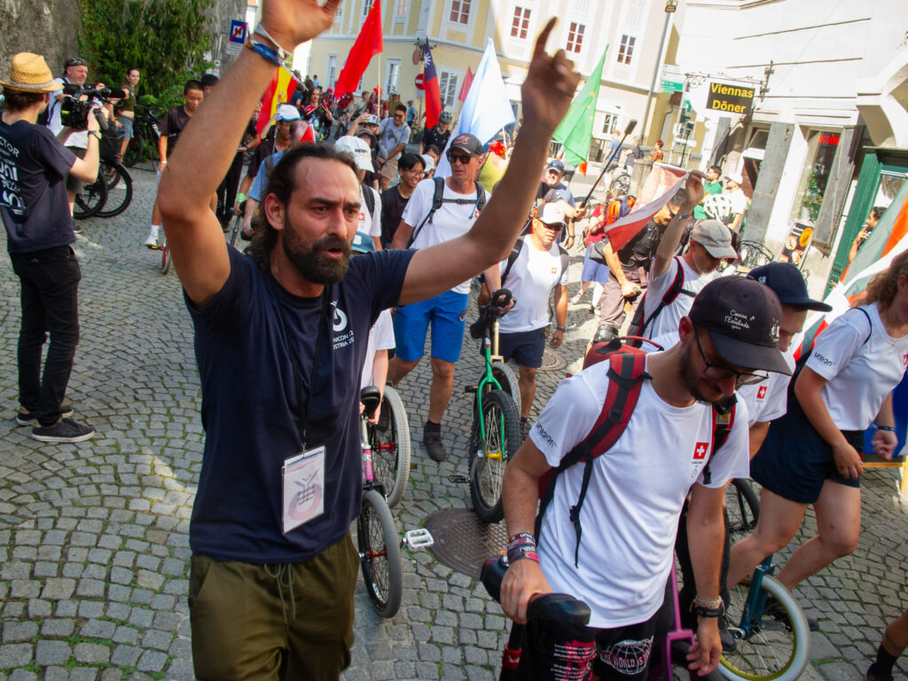 A crowd of unicyclists walks through a historic village carrying national flags. In the foreground, a man wearing a shirt that reads "UNICON 22, Austria 2026) raises his arms.