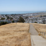 View from top of hill at McClaren Park in San Francisco. The hill in the foreground is grassy; a San Francisco neighborhood is in the background. A unicycle is climbing the trail.