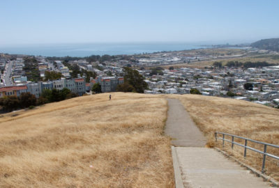 View from top of hill at McClaren Park in San Francisco. The hill in the foreground is grassy; a San Francisco neighborhood is in the background. A unicycle is climbing the trail.
