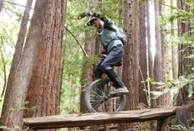 Man on mountain unicycle, riding on elevated skinny log through redwood forest