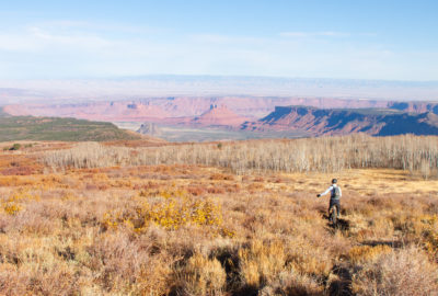A mountain unicyclist rides through an autumnal alpine landscape, with mostly bare aspens to the right, as the trail goes to the left towards a red rock desert with dramatic canyons and towers.