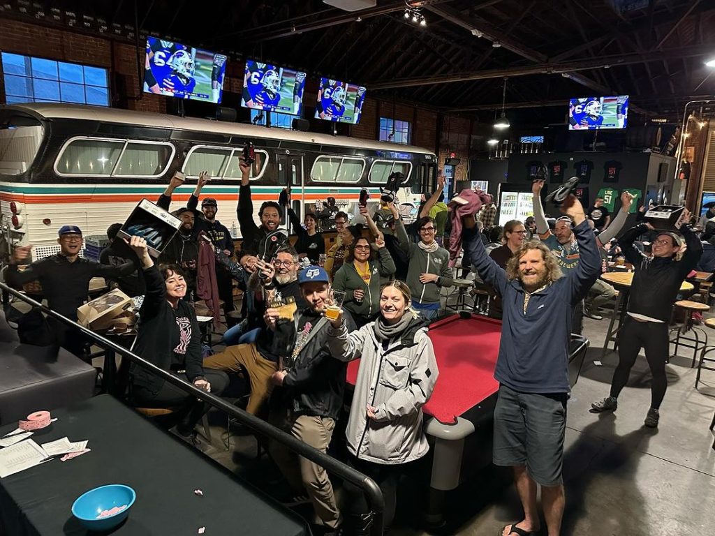 A group of people in a bar decorated by a classic AC Transit Bus raises their hands and glasses in triumph