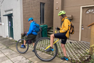 Two men prepare to mount unicycles with large wheels (29" and 36"). Both are poised with one foot on the ground and one on their left pedal. They wear athletic gear designed for light rain; one is in blue, the other in yellow.