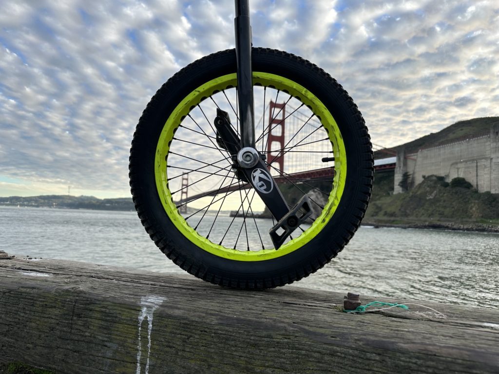 A BMX-style 19" unicycle wheel is held up in front of an afternoon sky with puffy clouds at the Golden Gate Bridge
