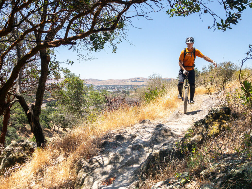 A mountain unicyclist rides on a rocky trail in the sun. The hill drops away behind him, revealing a valley and hills in the distance. He is approaching an area with many chunky rocks, shaded by oak trees.
