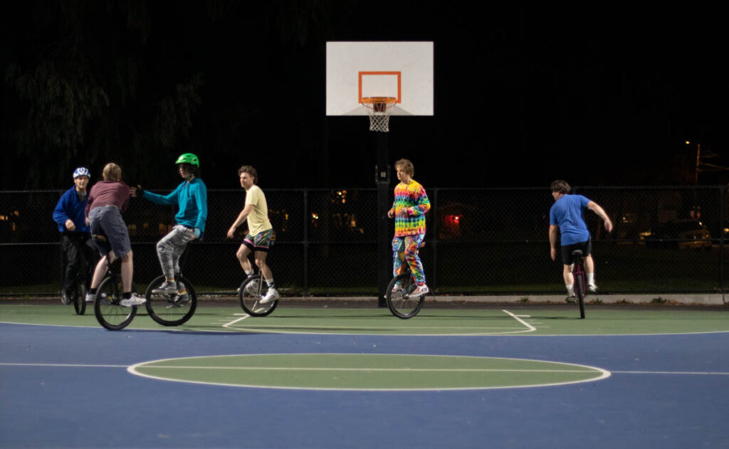 Five people on unicycles are seen on an outdoor basketball court at night. One is dribbling, with an apparent teammate cutting towards the basket on the other side of the image. A man in bright tie-dye is positioned in a zone defense position under the basket.