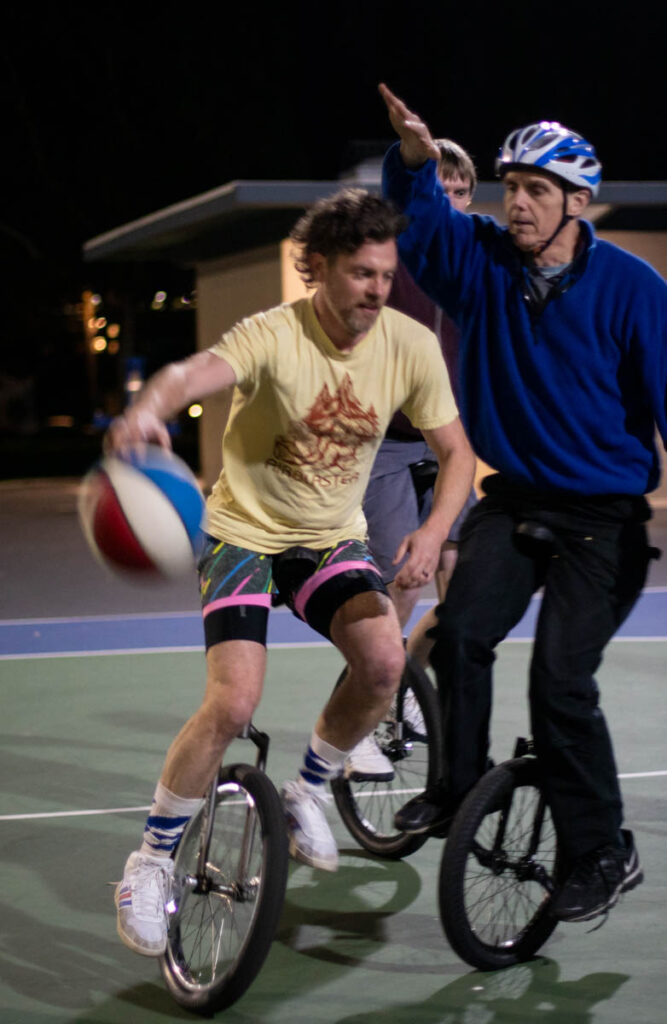 A man on a unicycling dribbles a basketball on an outdoor court at night. A taller man, also on a unicycle, is defending him with arms raised