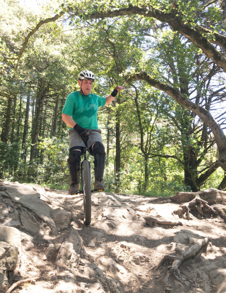 A mountain unicyclist, shirt soaked in sweat, rides down a steep rocky trail. His left arm is moving for balance, while his right tightly holds onto the unicycle's seat handle. He is concentrating intently.
