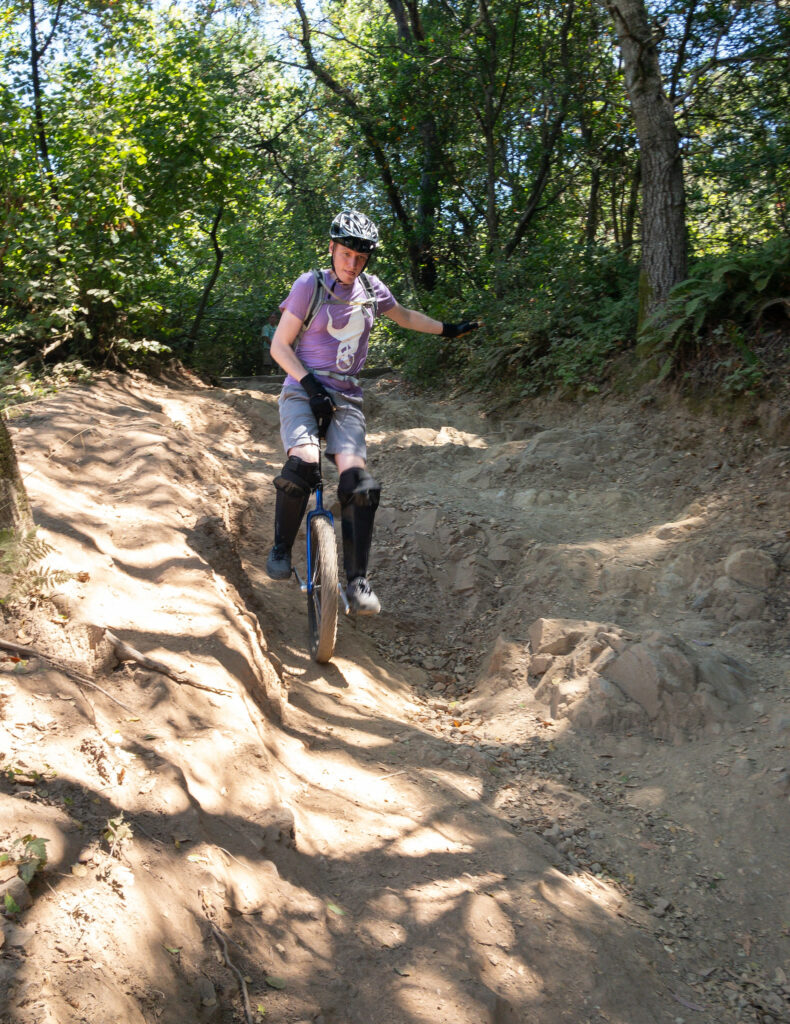 A mountain unicyclist rides down a steep, rocky trail in a forest. Sun dapples the trail ahead of him through the trees.