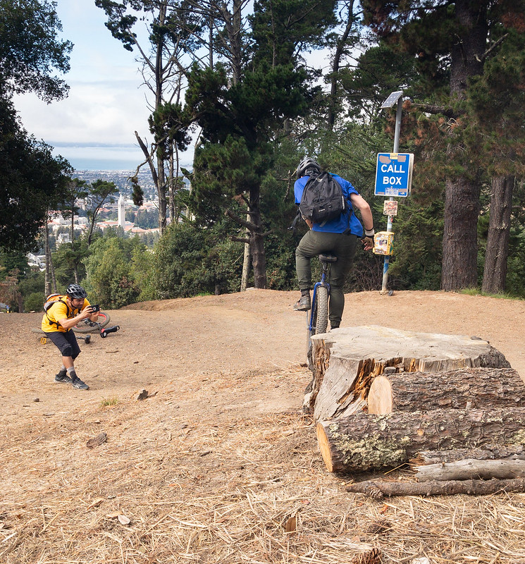 A mountain unicyclist drops off a large stump, about 2 feet high. A man in cycling gear crouches down in front of him with a camera. In the background is a stand of tall trees, with a gap revealing a view of Berkeley's Campanile and the San Francisco Bay.