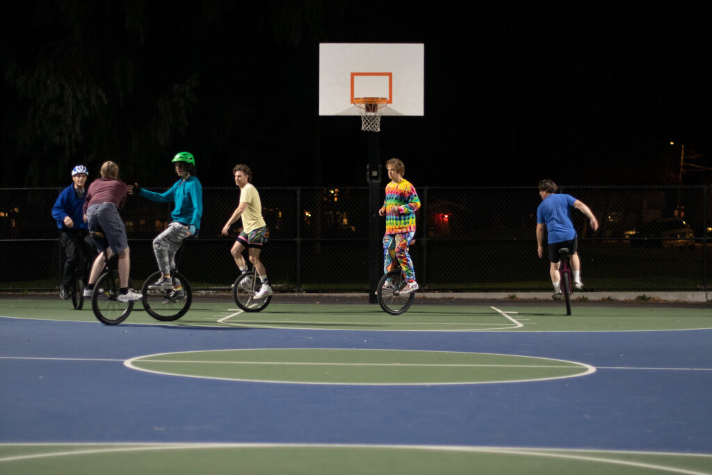 A group of five unicyclists play basketball on an outdoor court at night. The ball handler is on the left of the image near the three-point line, being defended by a tall man in blue. A man in a tie-dye shirt appears to be playing a zone defense under the basket, with another man in blue cutting towards the basket on the right side of the image.